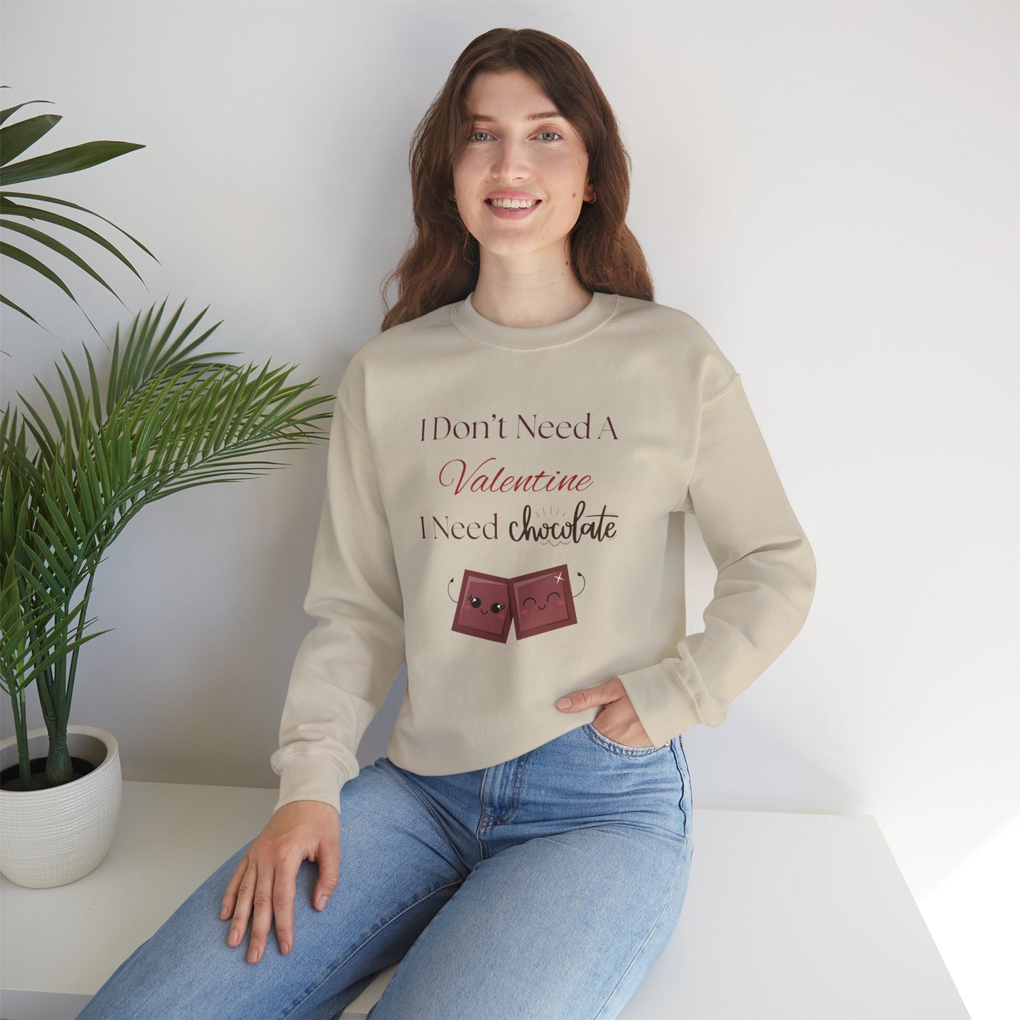 A woman seated wearing a tan Valentine’s sweatshirt that says “I Don’t Need a Valentine, I Need Chocolate,” with two cute smiling chocolate characters printed on the front.