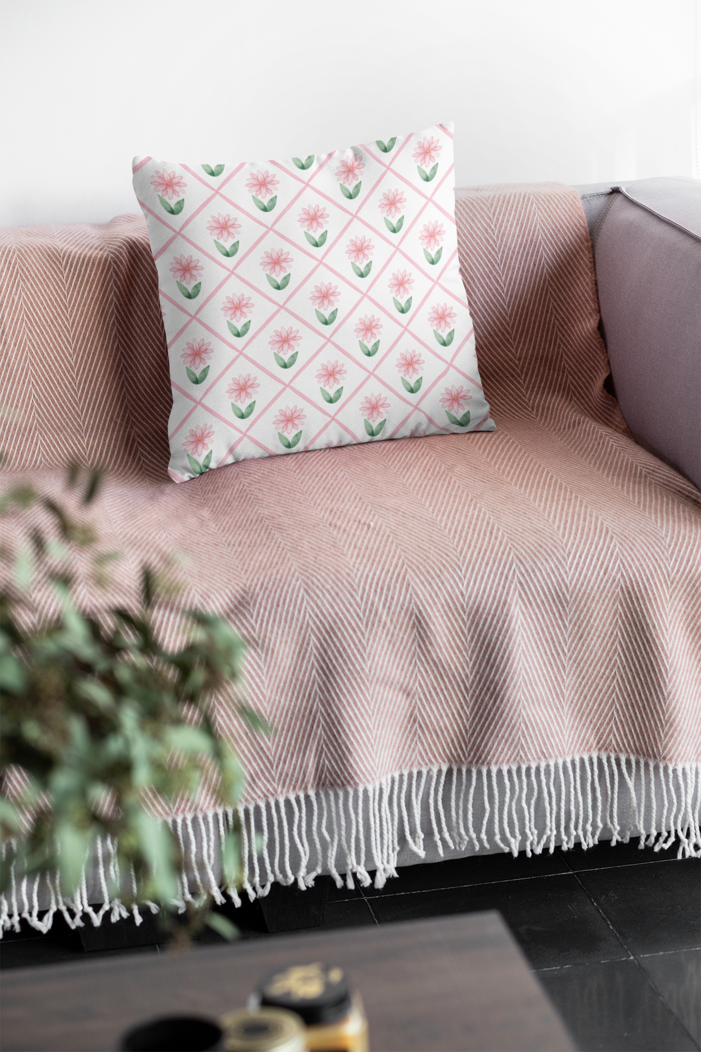 A soft pink daisy-patterned throw pillow with insert resting on a blush herringbone blanket on a cozy sofa.