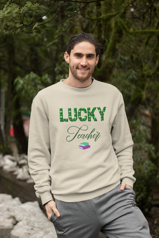 A smiling man wearing a cream-colored “Lucky Teacher” St. Patrick’s Day sweatshirt with green clover lettering, standing outdoors in a cozy forest-setting.