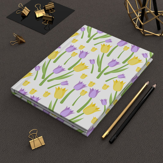 Hardcover journal with a repeating yellow and lavender tulip pattern, styled on a dark desk with gold binder clips and pencils.