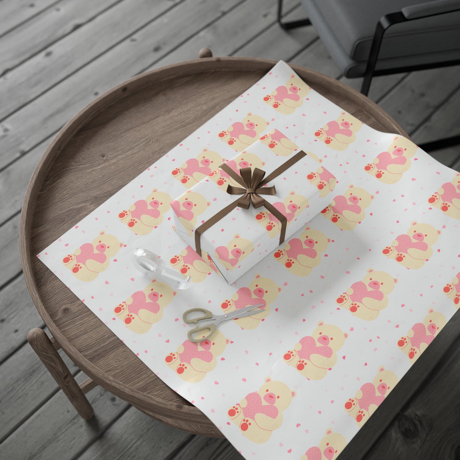 Cute teddy bear wrapping paper with pink hearts, displayed with a wrapped gift, scissors, and tape on a round wooden table.